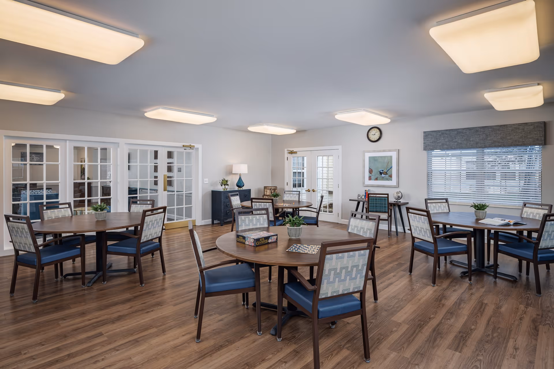 Bright senior living common dining room with round wooden tables, patterned chairs, potted plants, and large windows.
