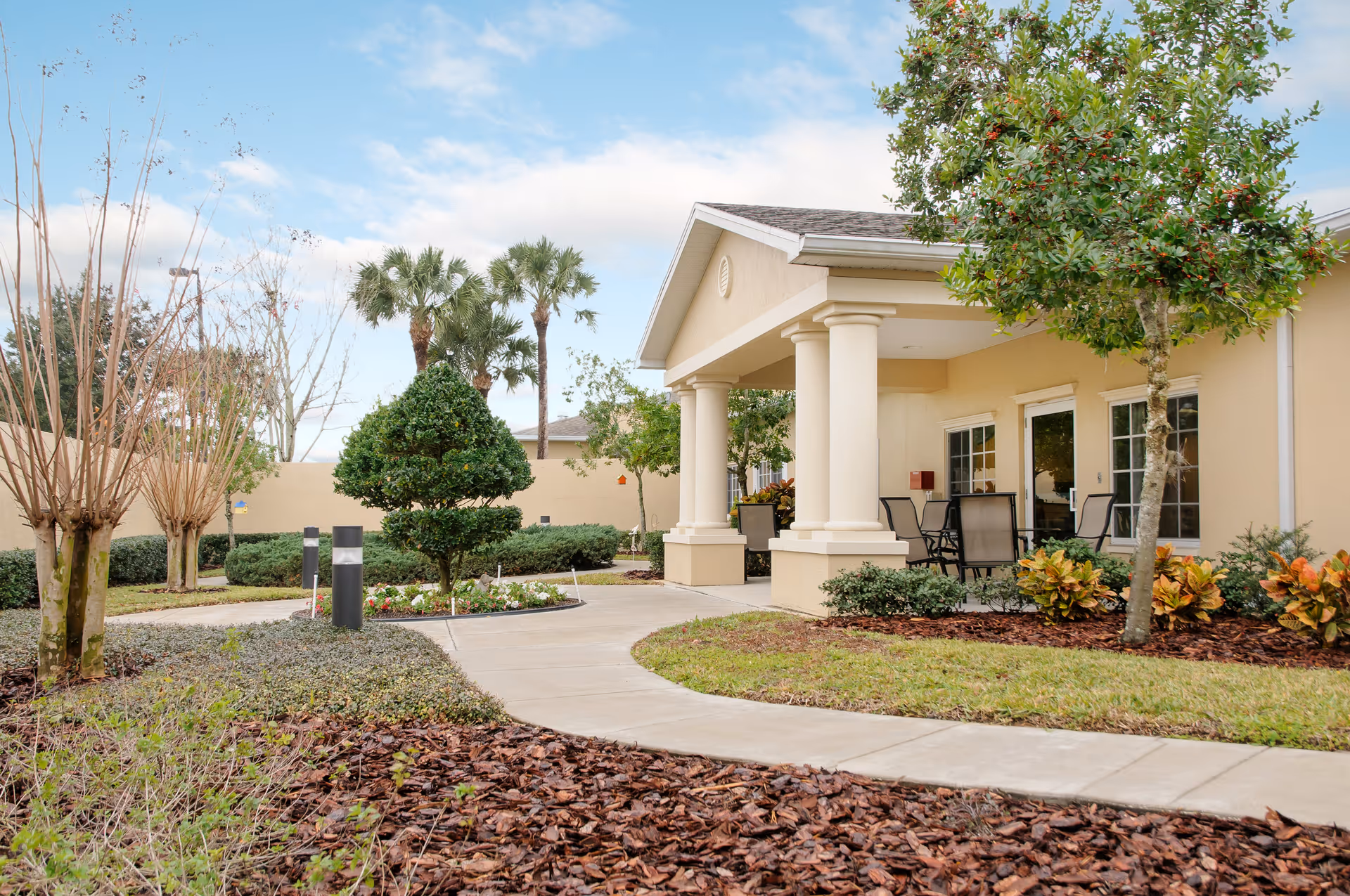 Front entrance of a single-story senior living facility with a covered portico, columns, outdoor seating, a curved walkway, and landscaped grounds.
