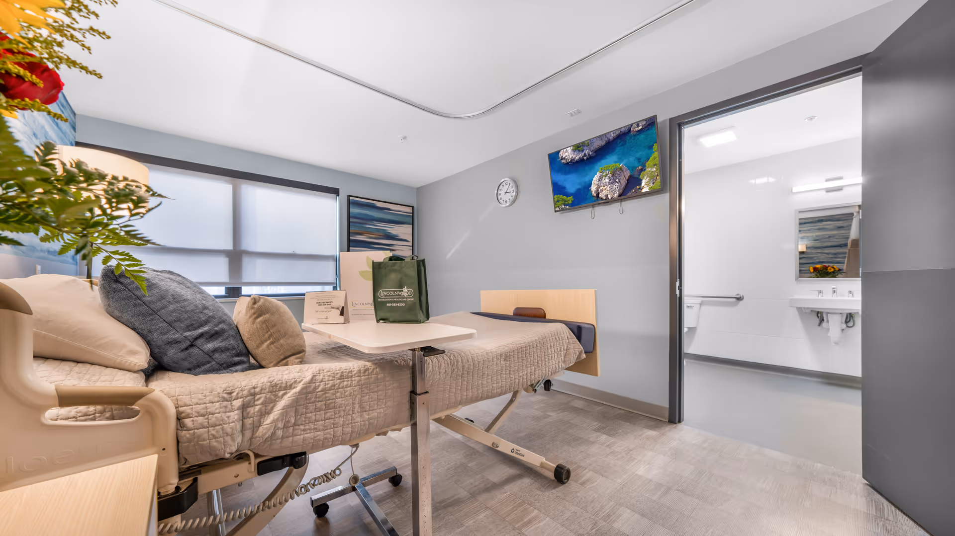 A bright and clean rehabilitation room with a hospital bed covered in beige bedding and multiple pillows. A small adjustable table is positioned over the bed. The room has a large window with frosted glass, a wall-mounted TV displaying a nature scene, a clock, and a door leading to a white-tiled bathroom with a sink and mirror. There are decorative plants and artwork on the walls.