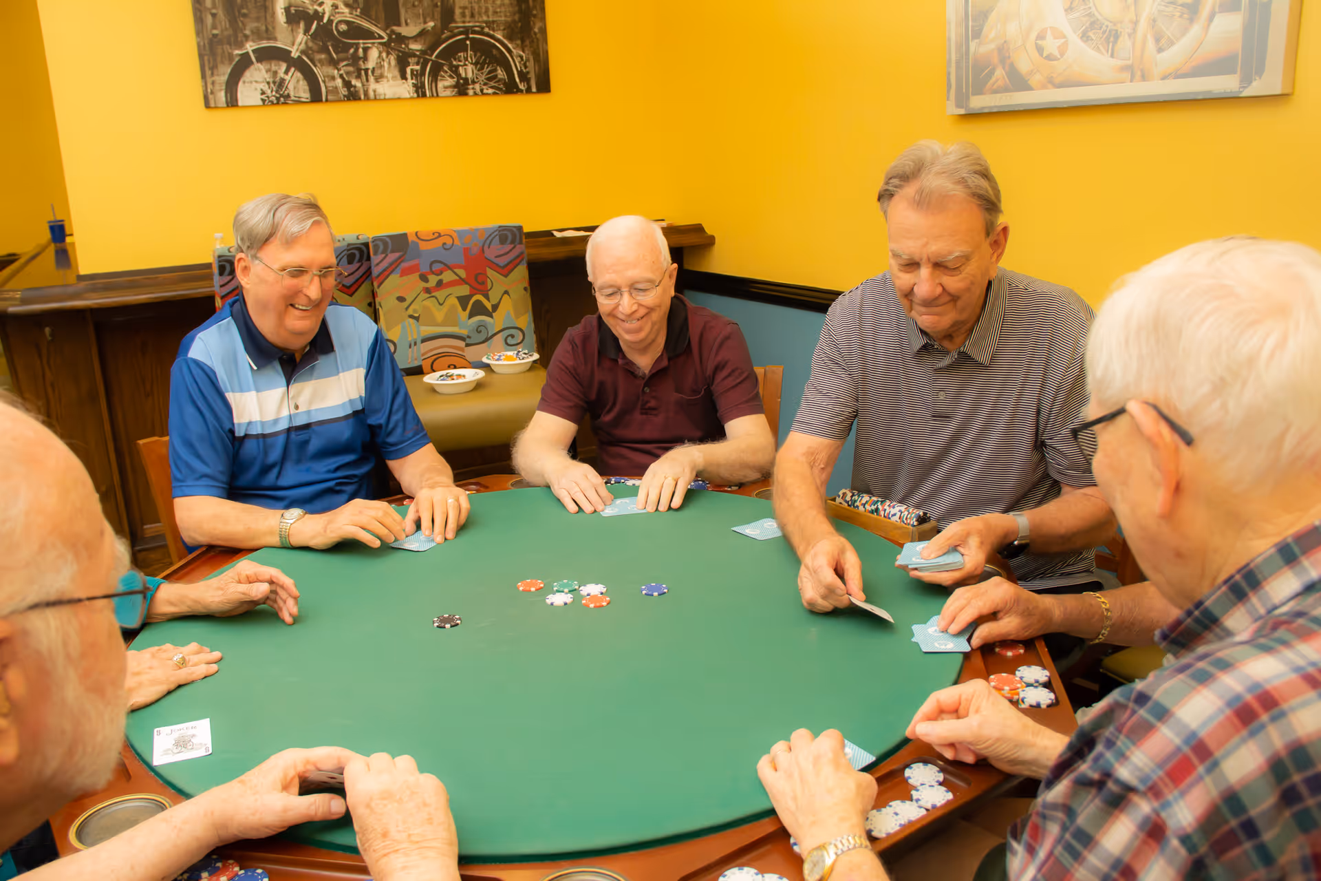 A group of six elderly men sitting around a green poker table playing cards and poker chips in a room with yellow walls and colorful chairs.