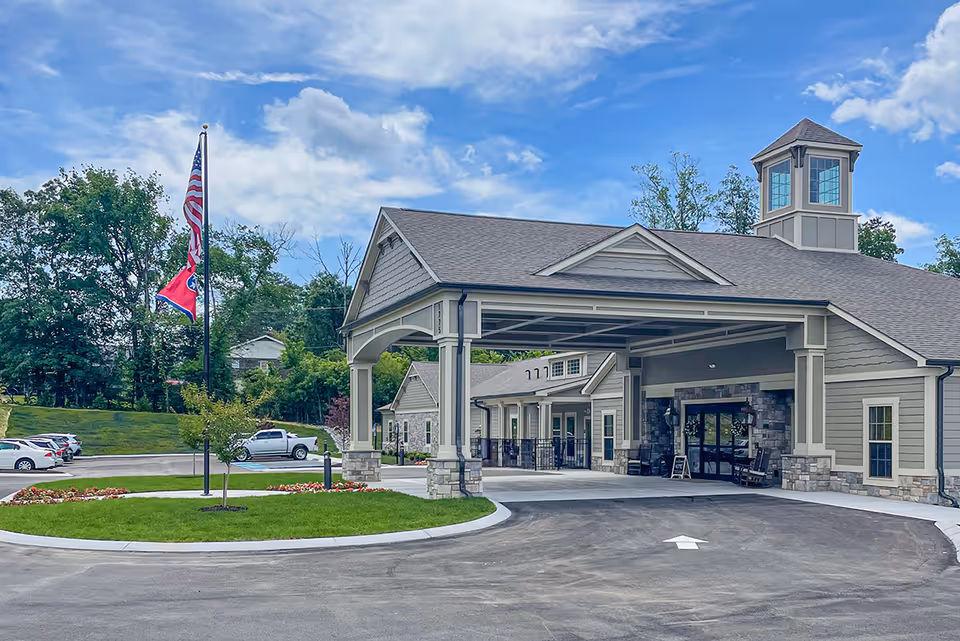 Exterior view of The Lantern at Morning Pointe Alzheimer's Center of Excellence, East Hamilton, showing a covered entrance with stone and siding facade, a driveway with an arrow, a flagpole with American and Tennessee state flags, and a parking area with several cars. The sky is partly cloudy.