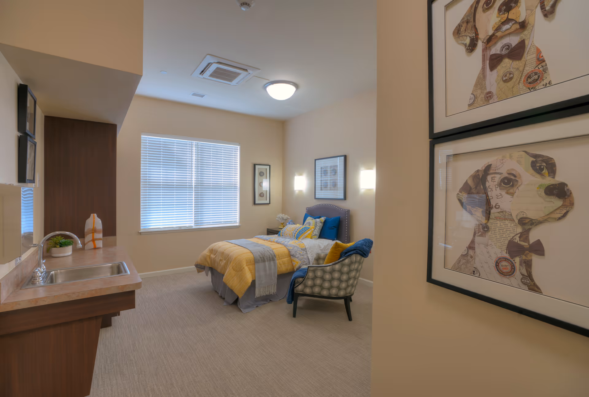 A cozy bedroom in a senior living facility with a single bed dressed in yellow and gray bedding, a patterned armchair with a blue throw, a window with blinds, and framed dog artwork on the wall near the entrance. There is also a small sink and countertop area on the left side of the room.