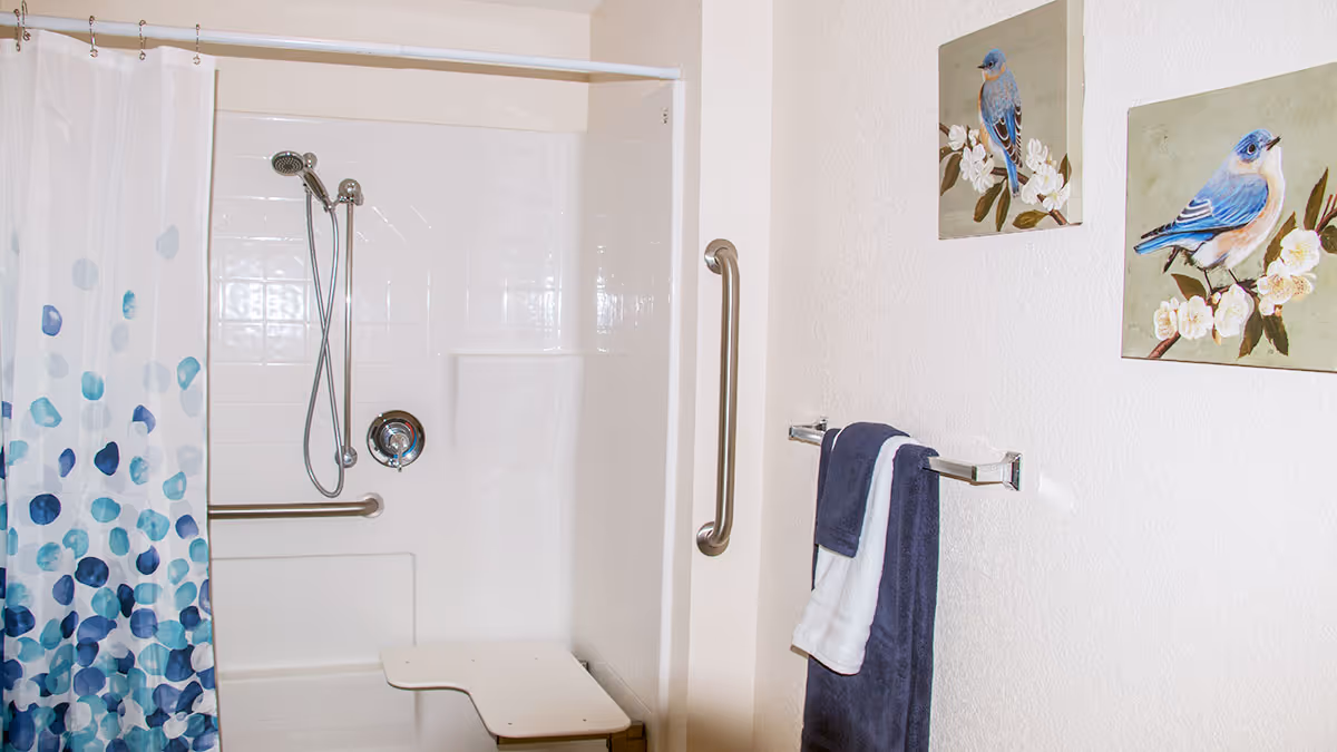 A bathroom shower area with a white tiled wall, a handheld showerhead, and a built-in seat. There is a white and blue patterned shower curtain on the left side. On the right wall, there are two framed paintings of blue birds on branches and a towel rack holding a dark blue and a white towel.
