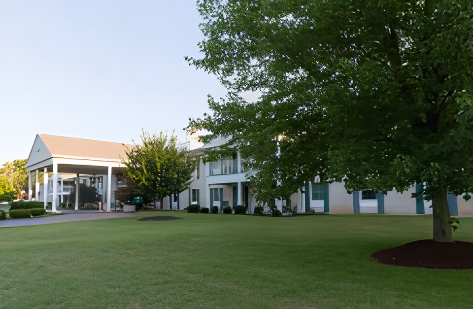Exterior view of a senior living facility building with a large green lawn and several trees in the foreground. The building has white walls, green window shutters, and a covered entrance area with columns.