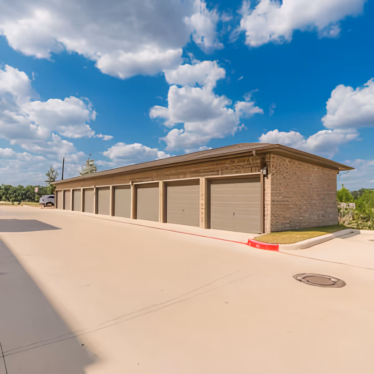 A row of closed garage doors attached to a single-story building under a partly cloudy blue sky, with a paved driveway in front and some greenery in the background.