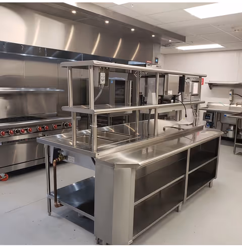 Commercial stainless-steel kitchen with a central prep island, shelving, and a large range under a ventilation hood.