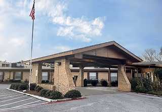 Exterior view of The Heights of Tyler senior living facility showing the front entrance with a covered drop-off area supported by stone pillars, an American flag on a flagpole, and a parking area in front under a partly cloudy sky.
