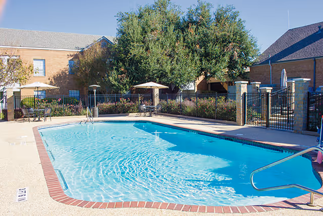 Outdoor swimming pool with clear blue water surrounded by a concrete deck with brick edging. There are several patio tables with umbrellas and chairs around the pool area, enclosed by a black metal fence. In the background, there are brick buildings and large green trees under a clear blue sky.