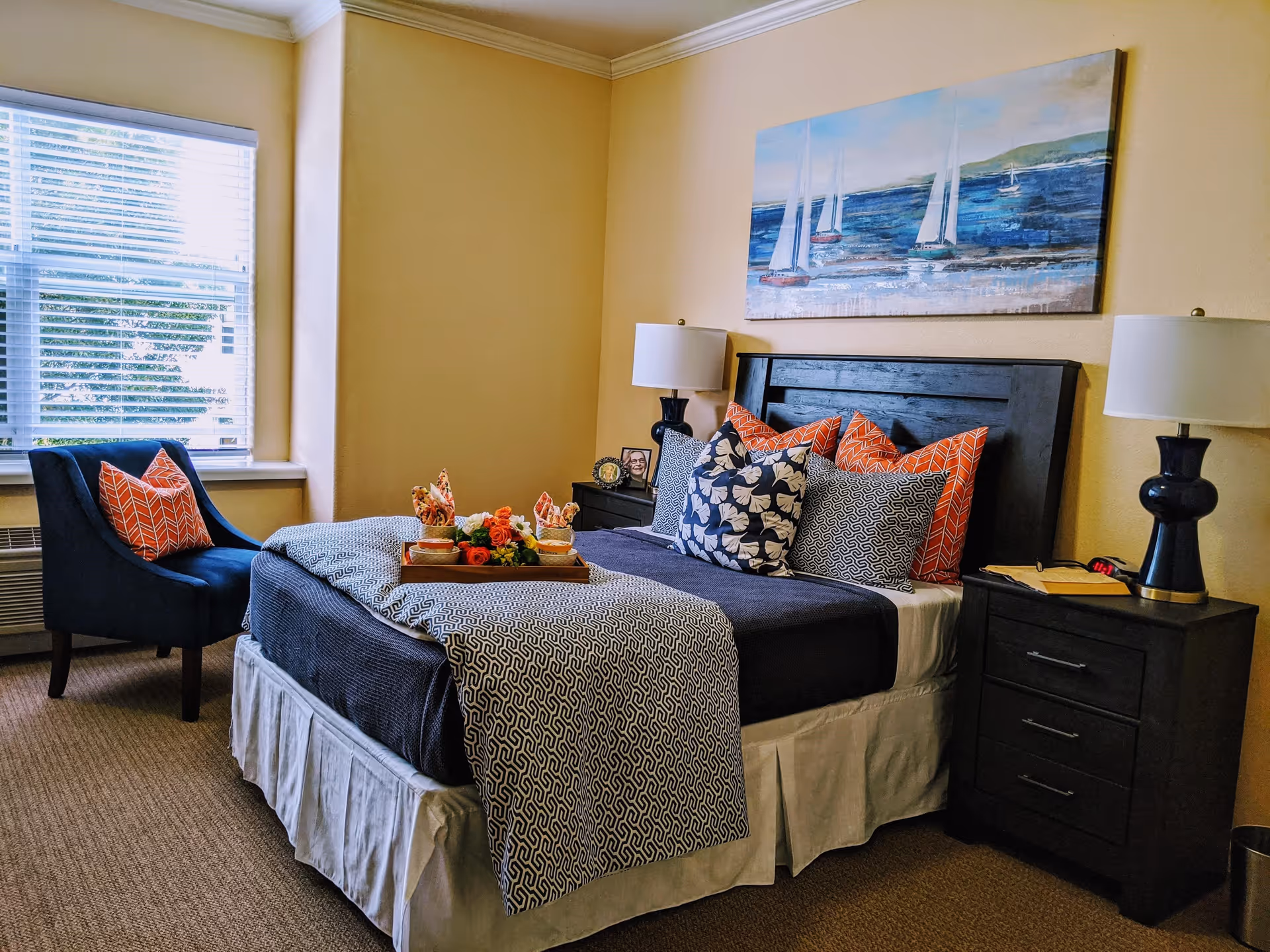A cozy bedroom with a neatly made bed featuring multiple decorative pillows in orange, black, and white patterns. There are two dark wooden nightstands on either side of the bed, each with a lamp. A tray with flowers and napkins is placed on the bed. A dark blue armchair with an orange pillow is positioned near a window with white blinds. Above the bed hangs a painting of sailboats on the water.