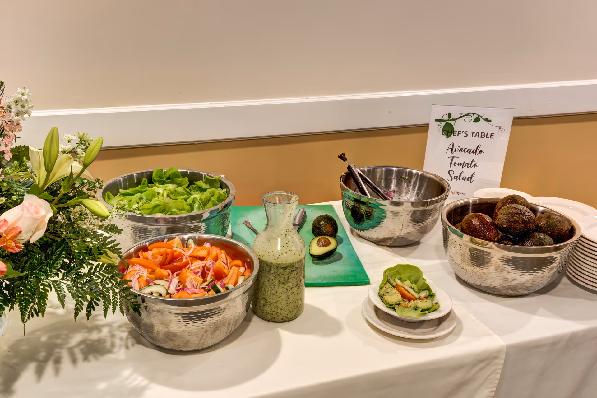 A buffet table with bowls of fresh salad ingredients including lettuce, sliced tomatoes, cucumbers, onions, and whole avocados. A bottle of green salad dressing is placed in the center. There is a small plate with a lettuce wrap containing avocado and tomato. A sign on the table reads 'Chef's Table Avocado Tomato Salad'. A floral arrangement is visible on the left side of the table.