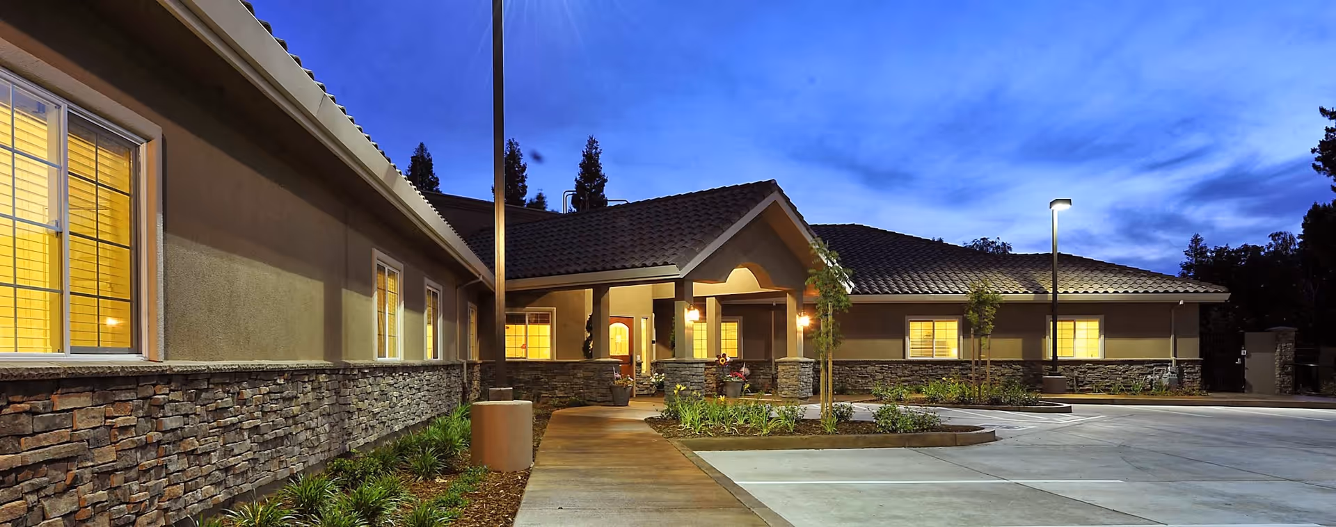 Exterior view of The Meadows at Country Place facility at dusk, showing a single-story building with stone and stucco facade, lit windows, a tiled roof, a walkway leading to the entrance, and a parking area with street lamps.