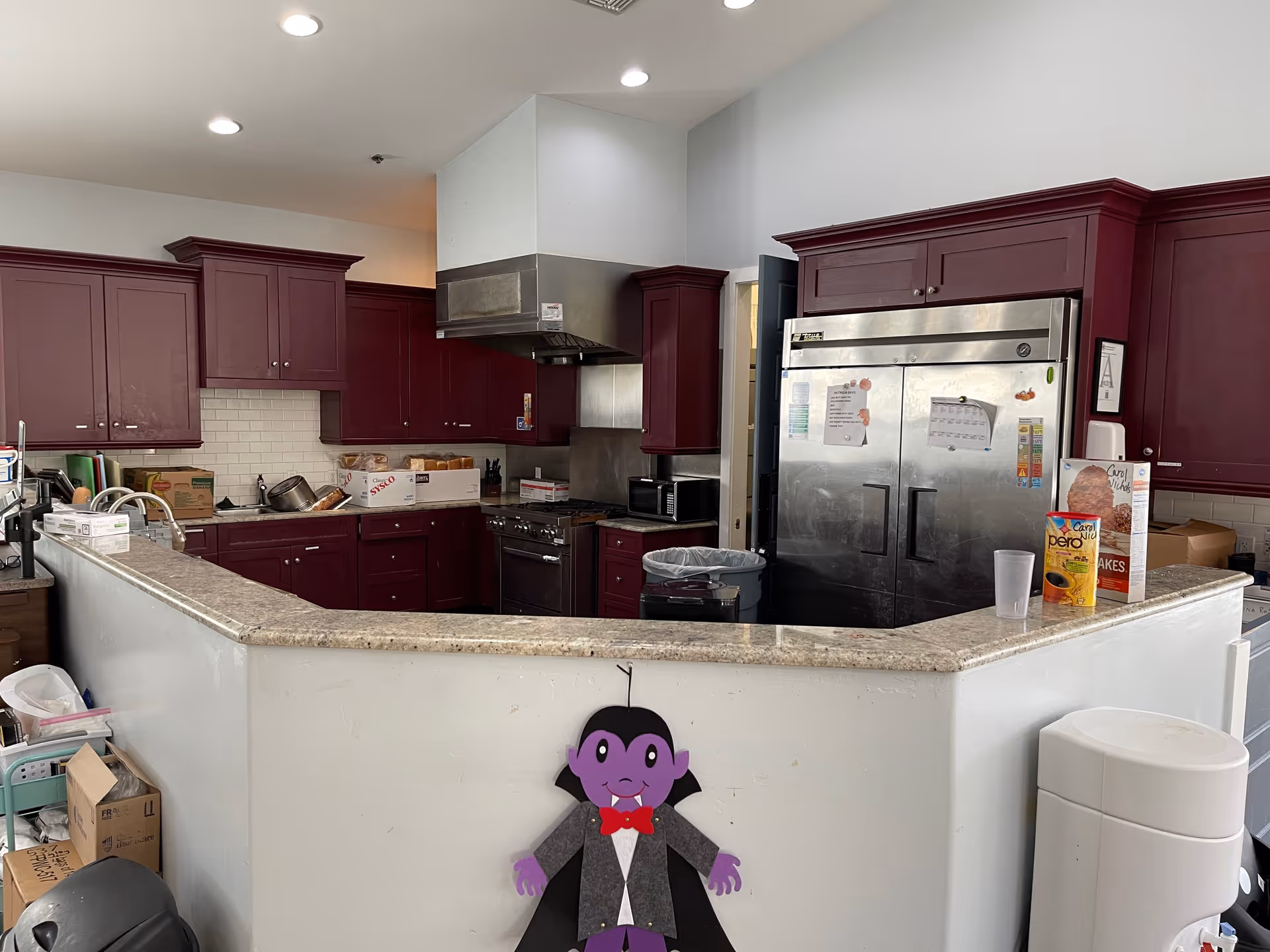 Commercial kitchen with burgundy cabinets, stainless steel refrigerator and range behind a curved serving counter decorated with a small purple vampire cutout.