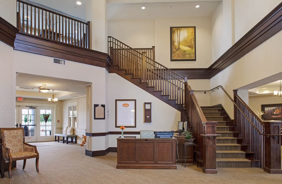 Lobby of a senior living facility with a wooden reception desk, seating area and a large staircase leading to an upper level.