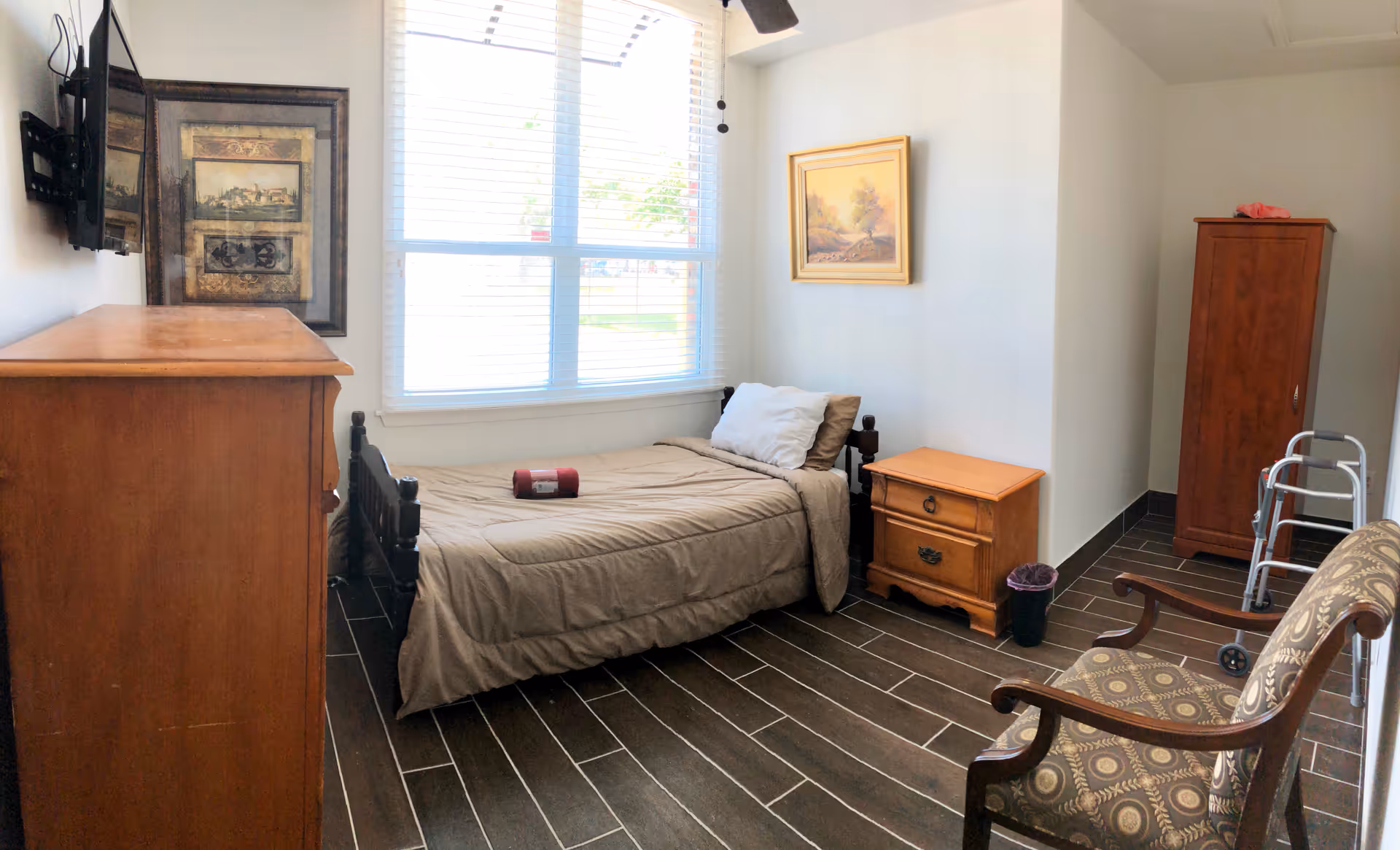 A small, neatly arranged bedroom in an assisted living facility featuring a single bed with beige bedding and pillows, a wooden nightstand with two drawers, a wooden dresser, a patterned armchair, a walker, a wooden wardrobe, and a large window with blinds letting in natural light. The walls are white, and there is framed artwork hanging on the walls.