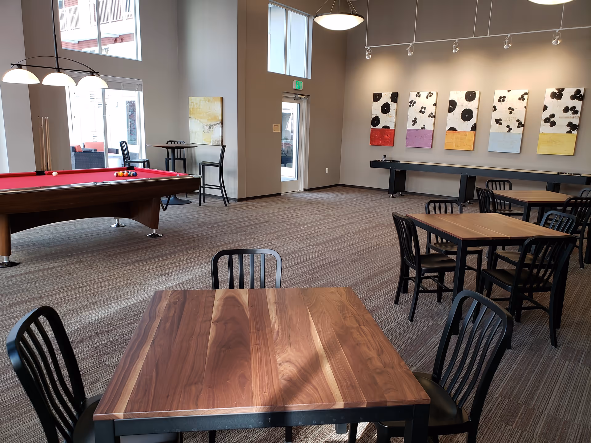 Spacious community activity room with a red-felt pool table, several wooden tables and black chairs, and colorful artwork on the wall.