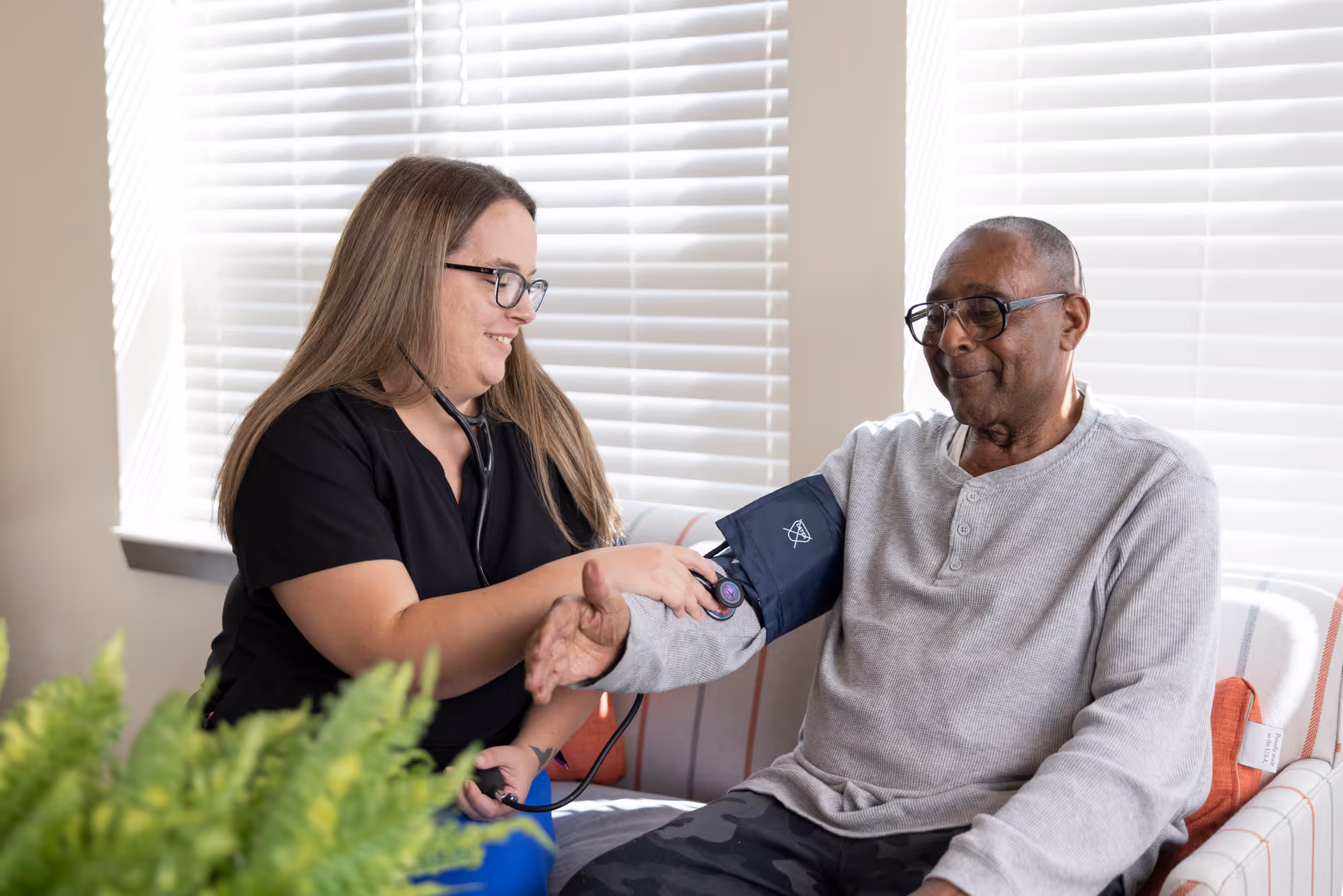 A healthcare professional wearing black scrubs and glasses is measuring the blood pressure of an elderly man sitting on a couch in a well-lit room with white blinds. The man is wearing a light gray long-sleeve shirt and glasses, and both appear calm and comfortable.
