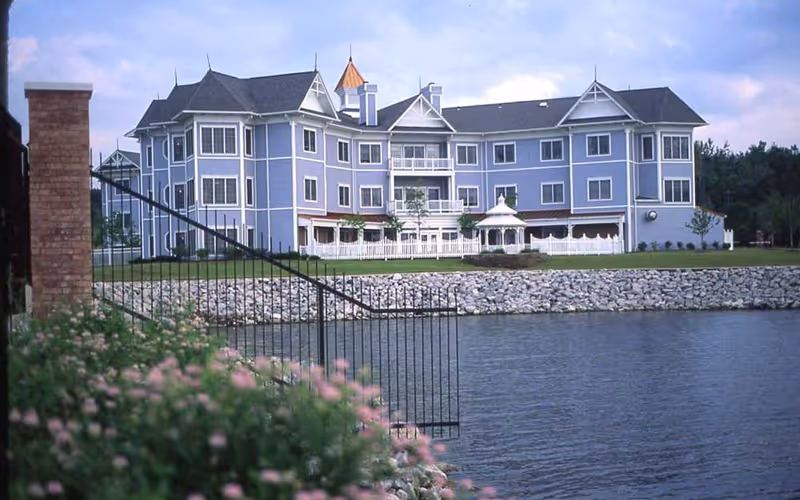 A large, multi-story residential building painted in light blue with white trim, situated beside a body of water. The building features multiple windows, balconies, and a small white gazebo in front. There is a stone embankment along the water and a black metal gate in the foreground with some pink flowers and greenery.