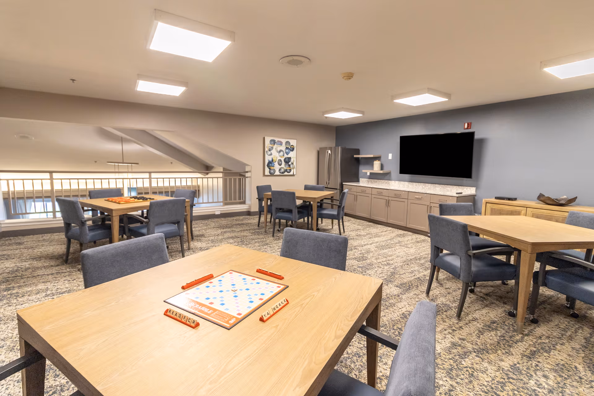 A spacious common area with multiple wooden tables and blue upholstered chairs arranged for group activities. One table in the foreground has a Scrabble board game set up. The room features a carpeted floor, a wall-mounted flat-screen TV, a refrigerator, cabinets, and shelves along one wall. The ceiling has several square recessed lights, and there is a railing overlooking a lower level.