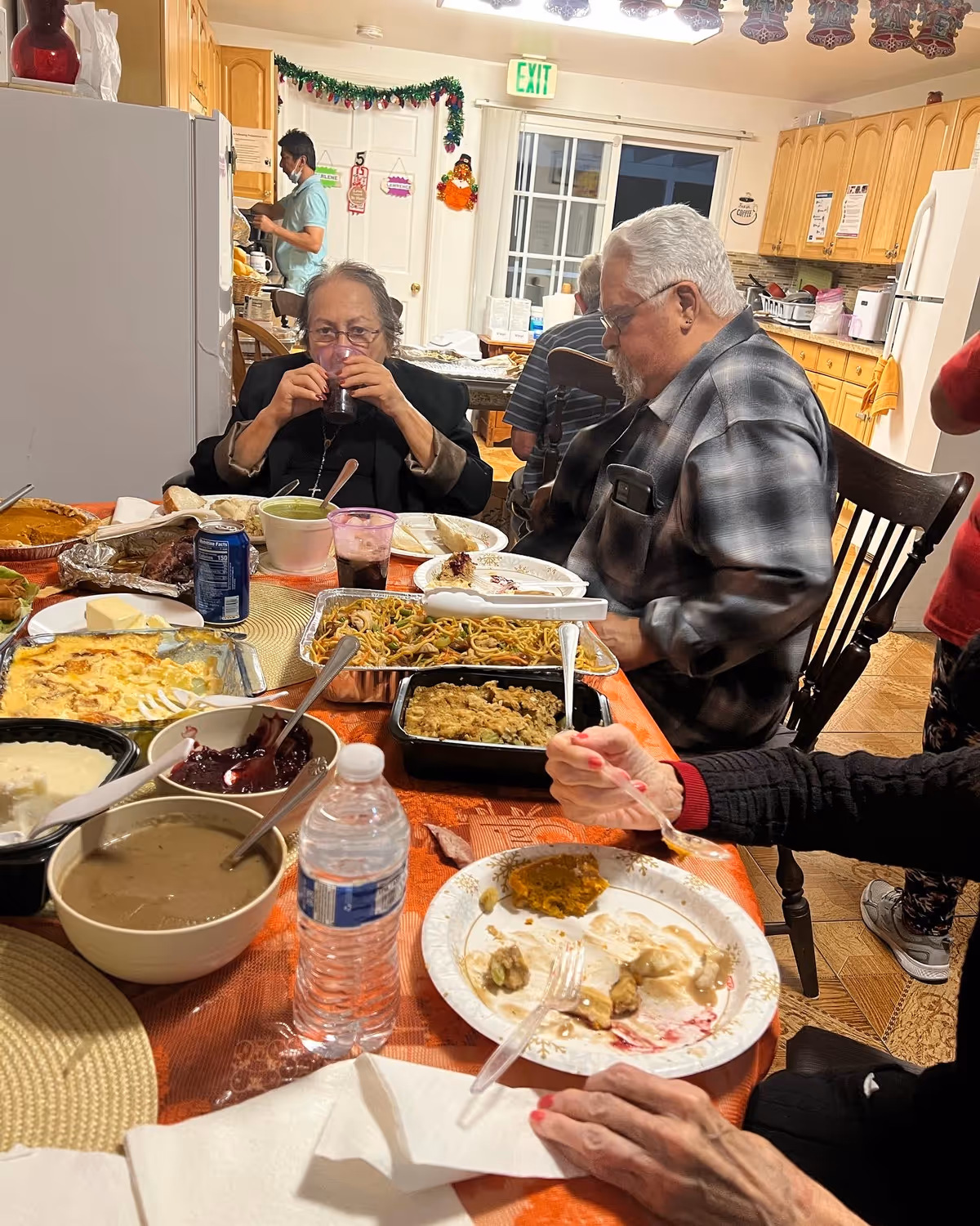 A group of elderly people sitting around a dining table filled with various dishes including noodles, mashed potatoes, gravy, cranberry sauce, and other food items. One woman is drinking from a glass, and another person is eating from a plate. In the background, a person wearing a mask is standing in a kitchen area with wooden cabinets and a refrigerator.