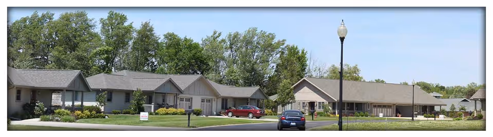 Front view of single-story senior living community buildings with landscaped lawns, parked cars, and trees under a clear sky.