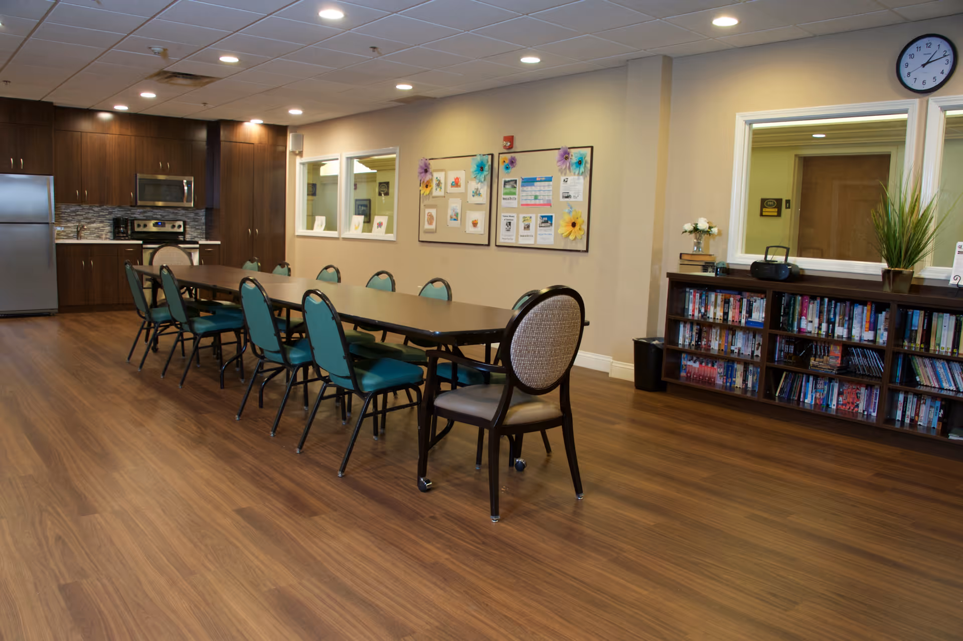 A senior living community common room with a long table and chairs, a kitchenette, bulletin board, and bookshelf.