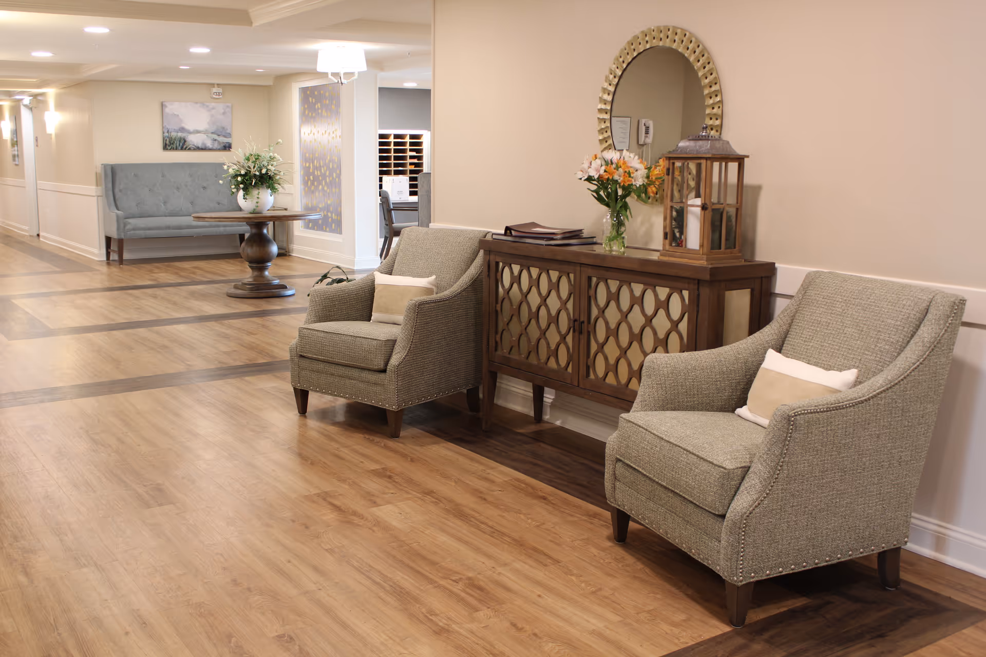 A well-lit hallway in a senior living facility with two upholstered armchairs with pillows, a wooden cabinet with decorative lattice doors, a round mirror, a lantern, and a vase of flowers on top. In the background, there is a round wooden table with a large floral arrangement and a blue upholstered bench against the wall.