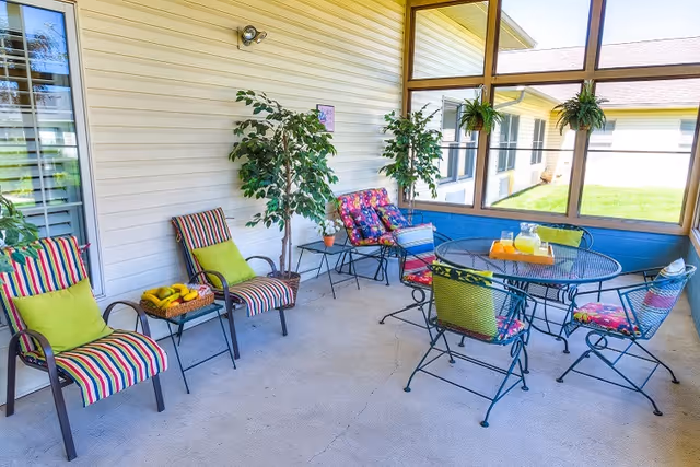 A bright and colorful screened-in porch with striped and floral cushioned chairs, green pillows, a round metal table with a pitcher of lemonade and glasses, potted plants, and a basket of fruit on a small side table.