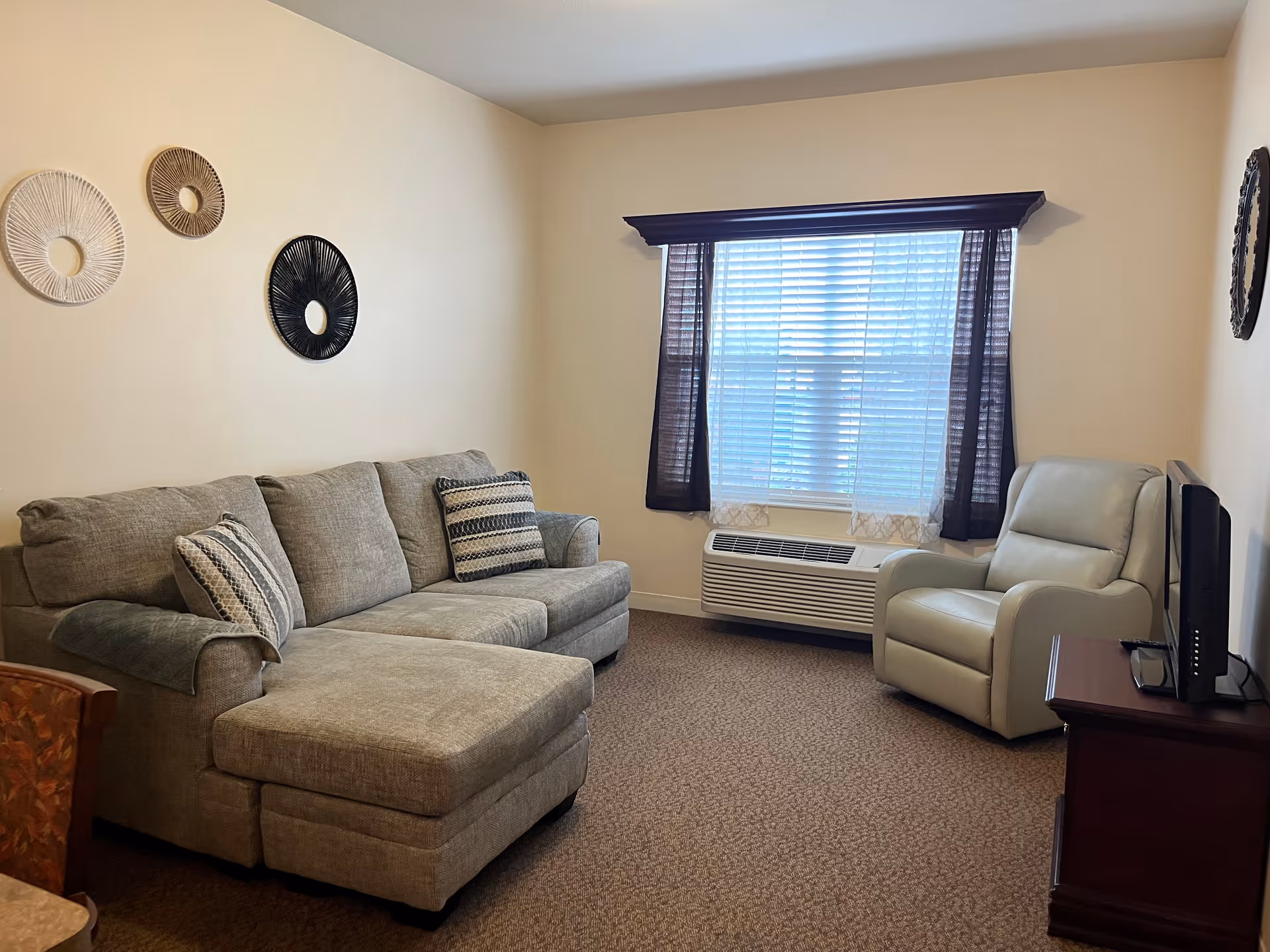 A cozy living room with a gray sectional sofa adorned with patterned pillows, a light-colored recliner chair, a window with blinds and dark curtains, a wall-mounted air conditioning unit below the window, and decorative circular wall hangings on the beige walls.