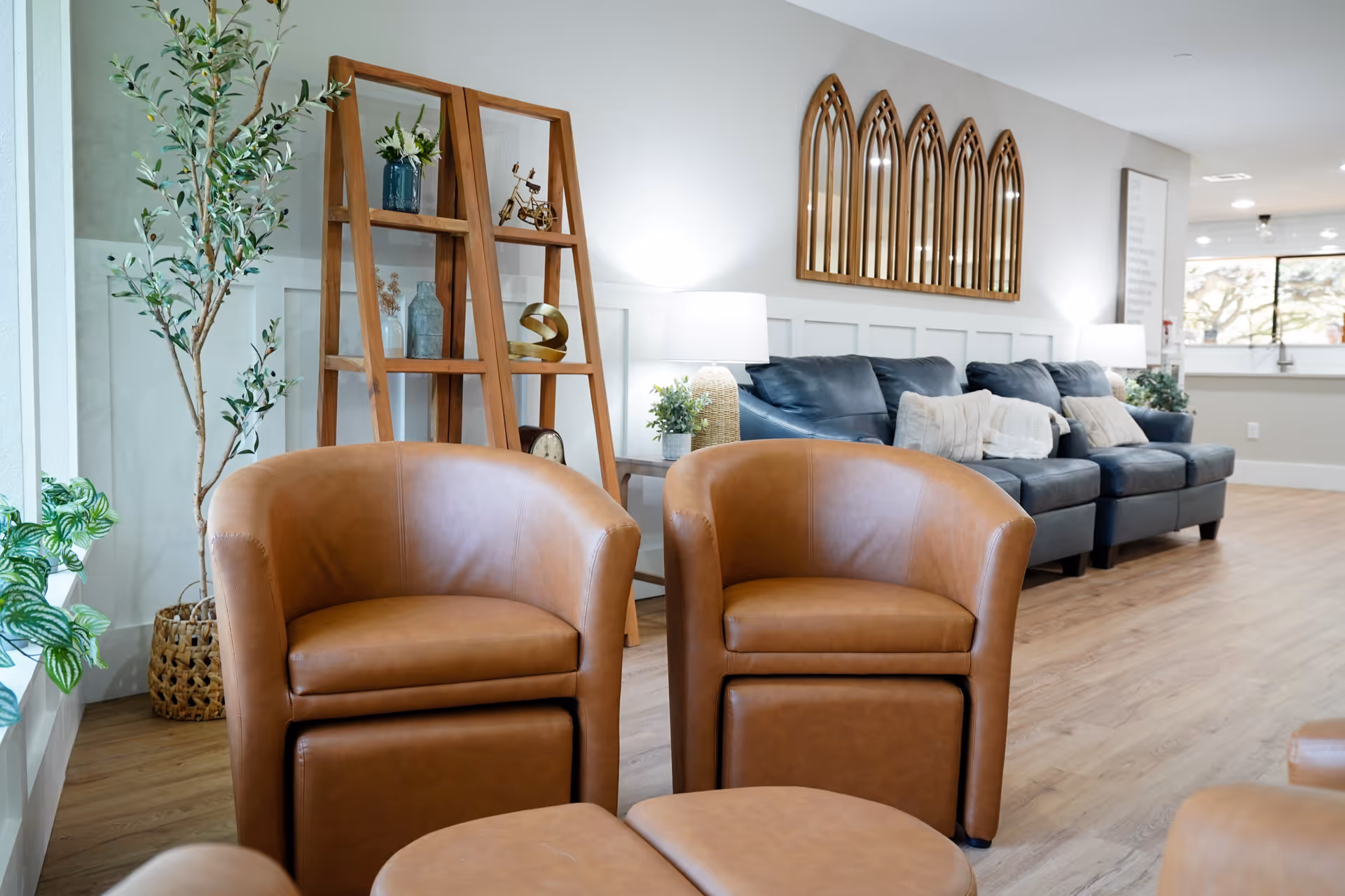 A bright and modern living room area with two brown leather armchairs in the foreground, a wooden shelving unit with decorative items, a tall potted plant, and a dark blue sofa with cushions against the wall. The room has wooden flooring, white walls, and decorative mirrors shaped like arched windows above the sofa.