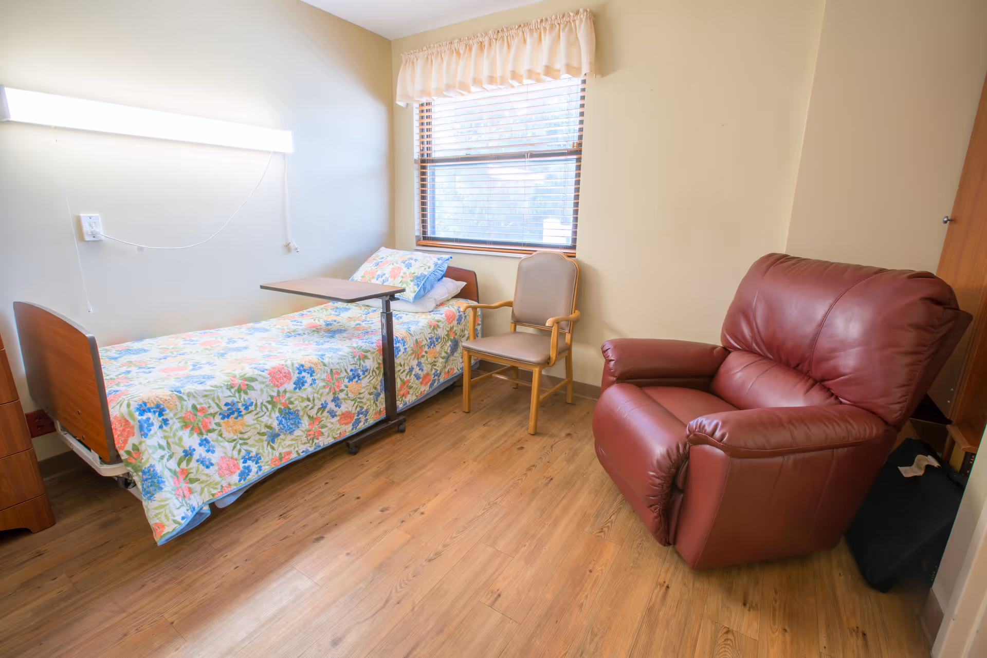 A cozy bedroom in a senior living facility with a hospital bed, a chair, and natural light from a window.