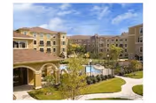Outdoor view of Evergreen at Plano senior living facility showing a multi-story building surrounding a courtyard with a swimming pool, landscaped greenery, and walking paths under a partly cloudy sky.