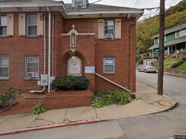 Exterior view of a red brick building on a street corner with a small statue niche above an arched plaque. There are windows with shutters and an air conditioning unit. A sidewalk and a street with a parked car and houses are visible.