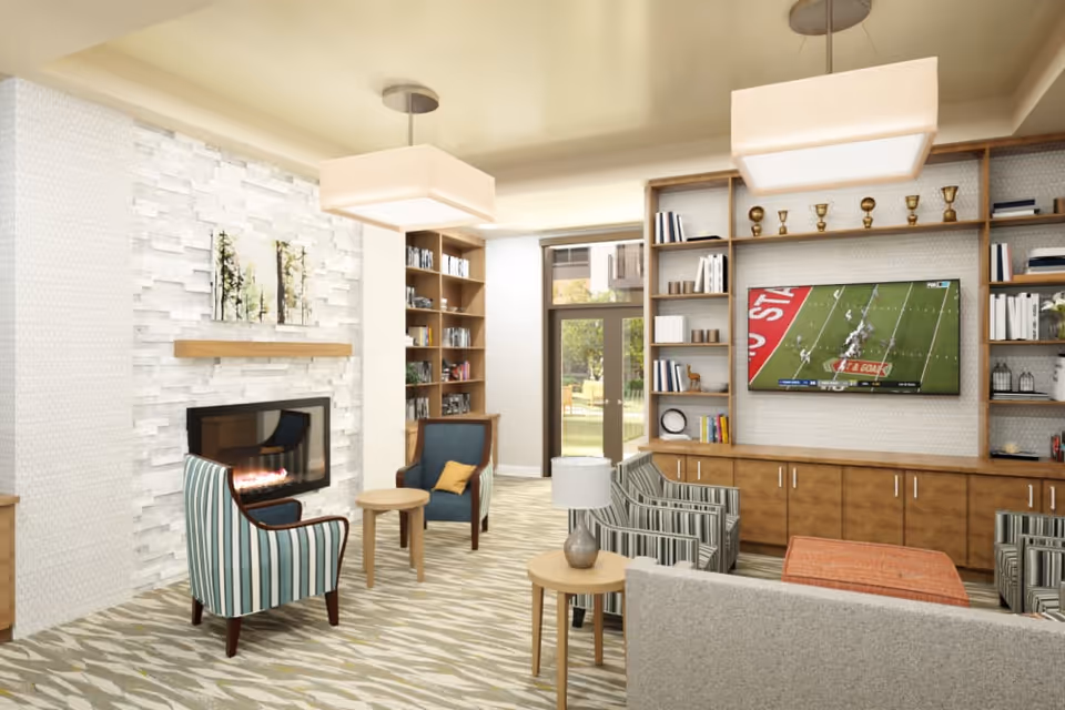 A cozy living room area with a modern fireplace set in a white textured wall. There are several armchairs with striped and solid upholstery arranged around small wooden tables. A large flat-screen TV mounted on a wooden built-in shelving unit displays a football game. The room has large windows letting in natural light and two rectangular ceiling lights.