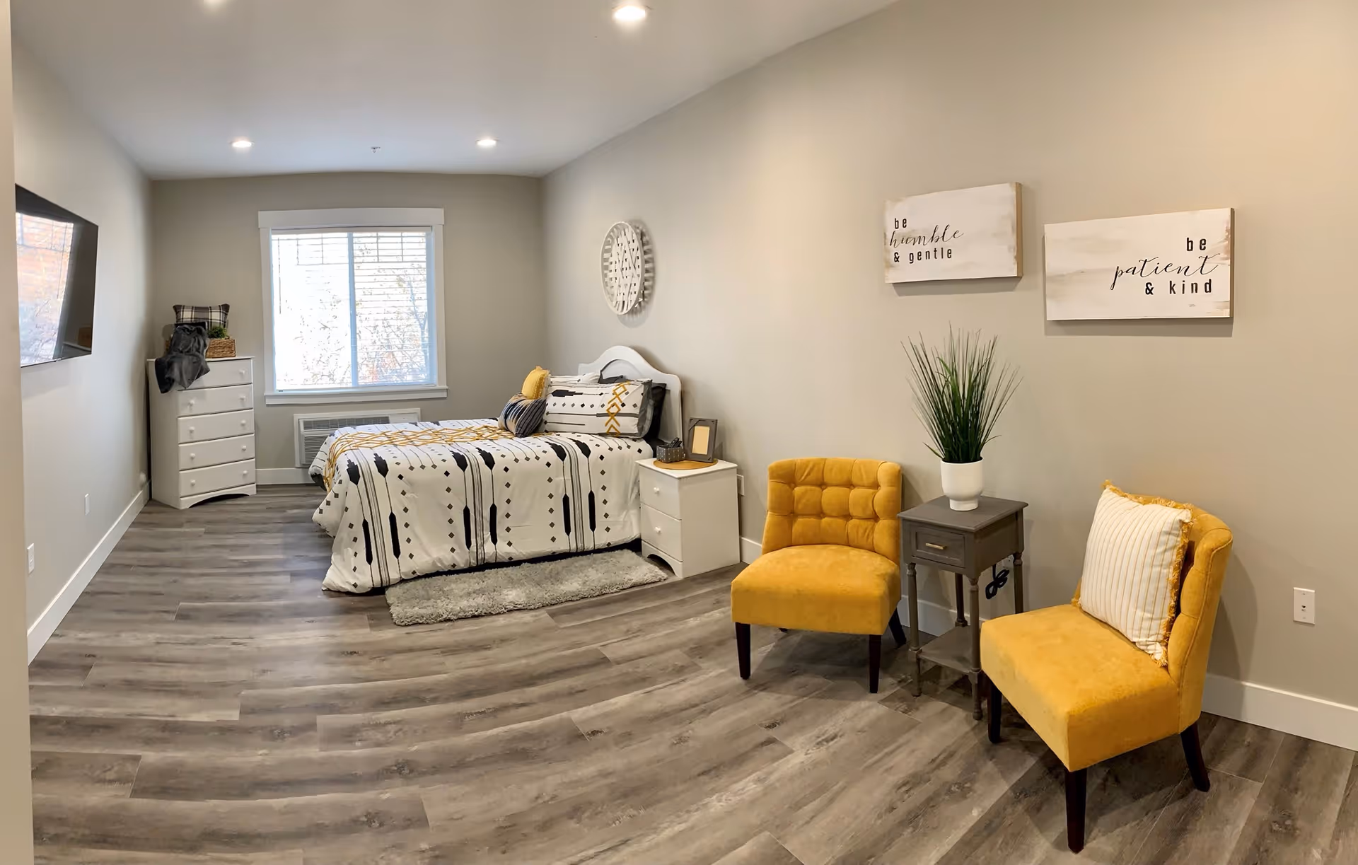 A bright and modern bedroom with a bed featuring a white and black patterned comforter with yellow accents. There is a white nightstand with a small picture frame and decorative items next to the bed. Two mustard yellow upholstered chairs with a small gray side table and a potted plant are positioned against the wall. The walls are light beige with two framed inspirational quotes hanging above the chairs. A large window with white blinds lets in natural light, and a flat-screen TV is mounted on the opposite wall. The floor is covered with wood-look vinyl planks.