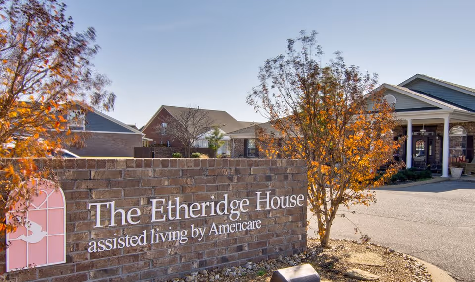 Exterior view of The Etheridge House assisted living facility showing a brick sign with the facility name and a building entrance in the background, surrounded by trees with autumn foliage.
