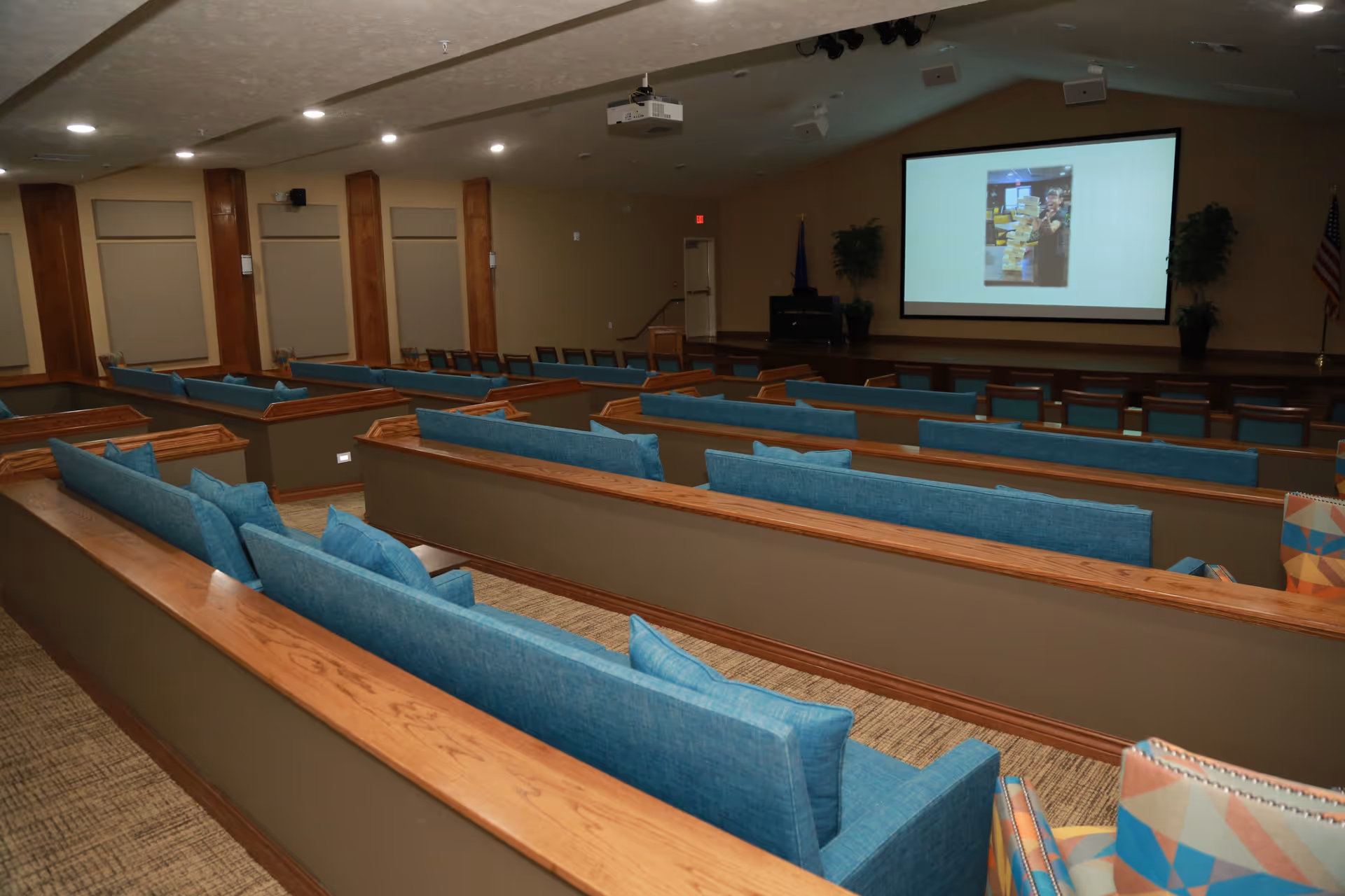 Interior view of a large room with multiple rows of blue cushioned pew-style seating facing a stage with a large projection screen displaying an image. The room has beige walls, wooden accents, and carpeted floors. There are potted plants on the stage and an American flag to the right.