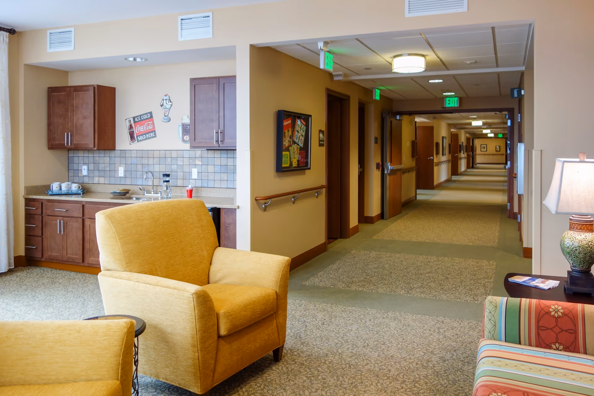 Interior view of a senior living facility lounge area with two yellow armchairs and a colorful striped sofa. There is a small kitchenette with wooden cabinets, a sink, and vintage-style wall decorations. A long hallway with multiple doors and exit signs extends into the background.