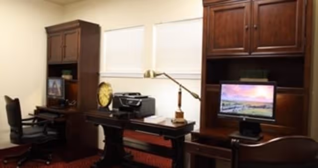 Interior office area with two dark wooden desks and matching chairs. One desk has a computer monitor displaying a scenic landscape, and the other desk has a printer and a large decorative clock. The room has beige walls and two windows with closed blinds.