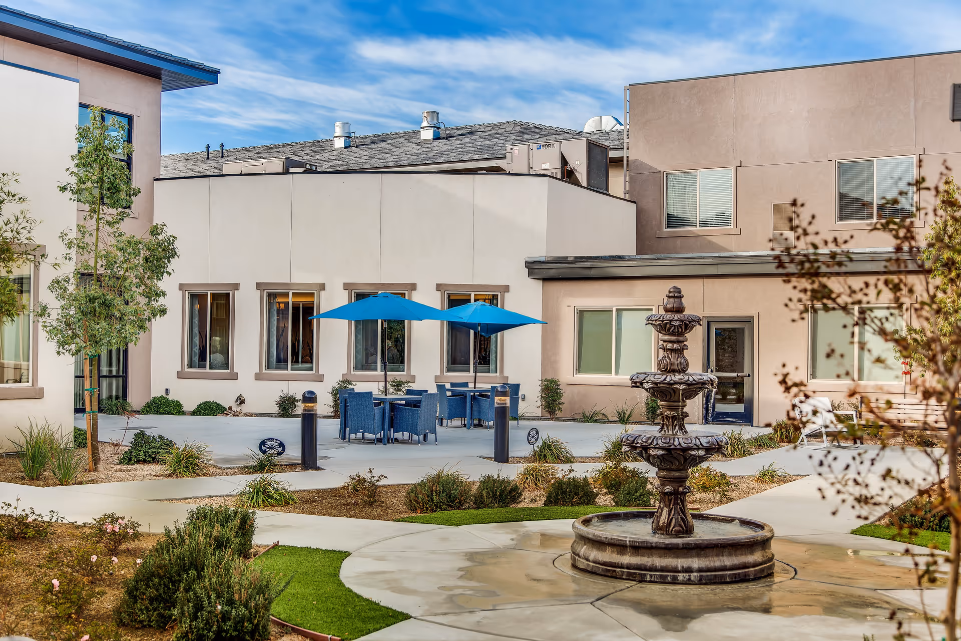 Outdoor courtyard area at Legacy House of Centennial Hills featuring a multi-tiered stone fountain in the foreground, landscaped garden beds with shrubs and small trees, and a seating area with blue chairs and tables shaded by blue umbrellas. The courtyard is surrounded by beige and light brown building walls with windows and a door, under a partly cloudy blue sky.