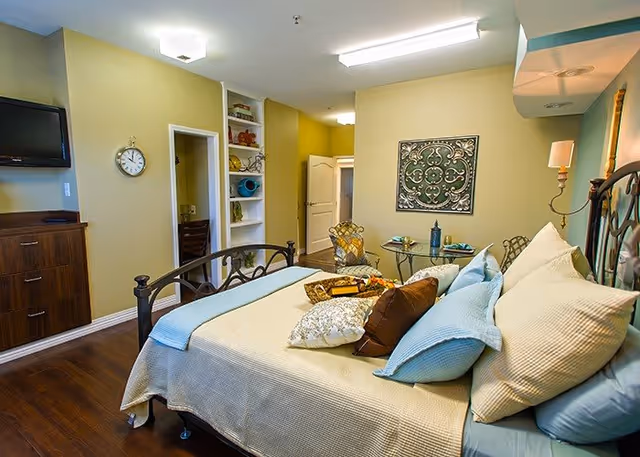 A cozy bedroom in a senior living facility with a neatly made bed featuring multiple pillows and a light blue throw blanket. The room has wooden flooring, a wall-mounted TV, a clock, built-in shelves with decorative items, a small round glass table with two chairs, and a decorative wall hanging. The walls are painted light green and there is a door leading to another room.