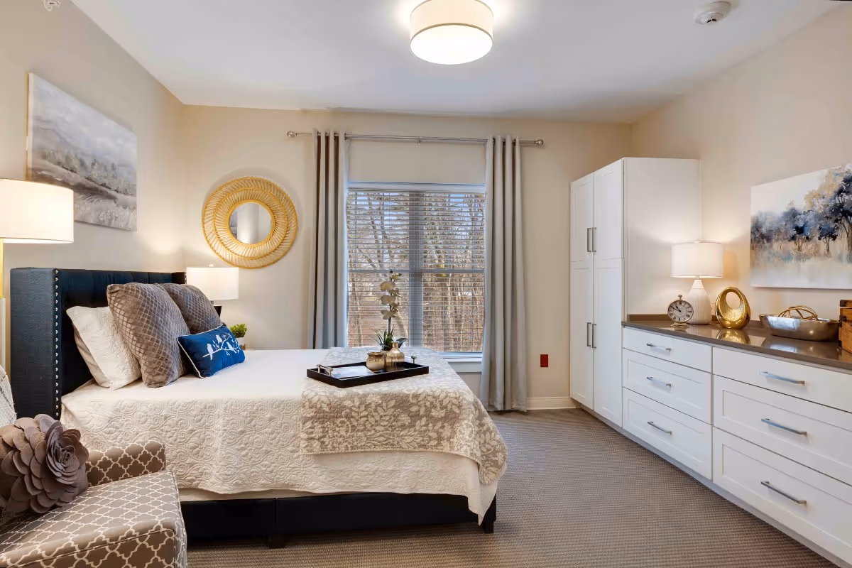 A well-lit bedroom featuring a neatly made bed with white and gray bedding, decorative pillows, and a tray with plants on top. There is a patterned armchair with a flower-shaped pillow, a round gold-framed mirror on the wall, a large window with gray curtains, white cabinetry with drawers and a countertop, two table lamps, and artwork on the walls.