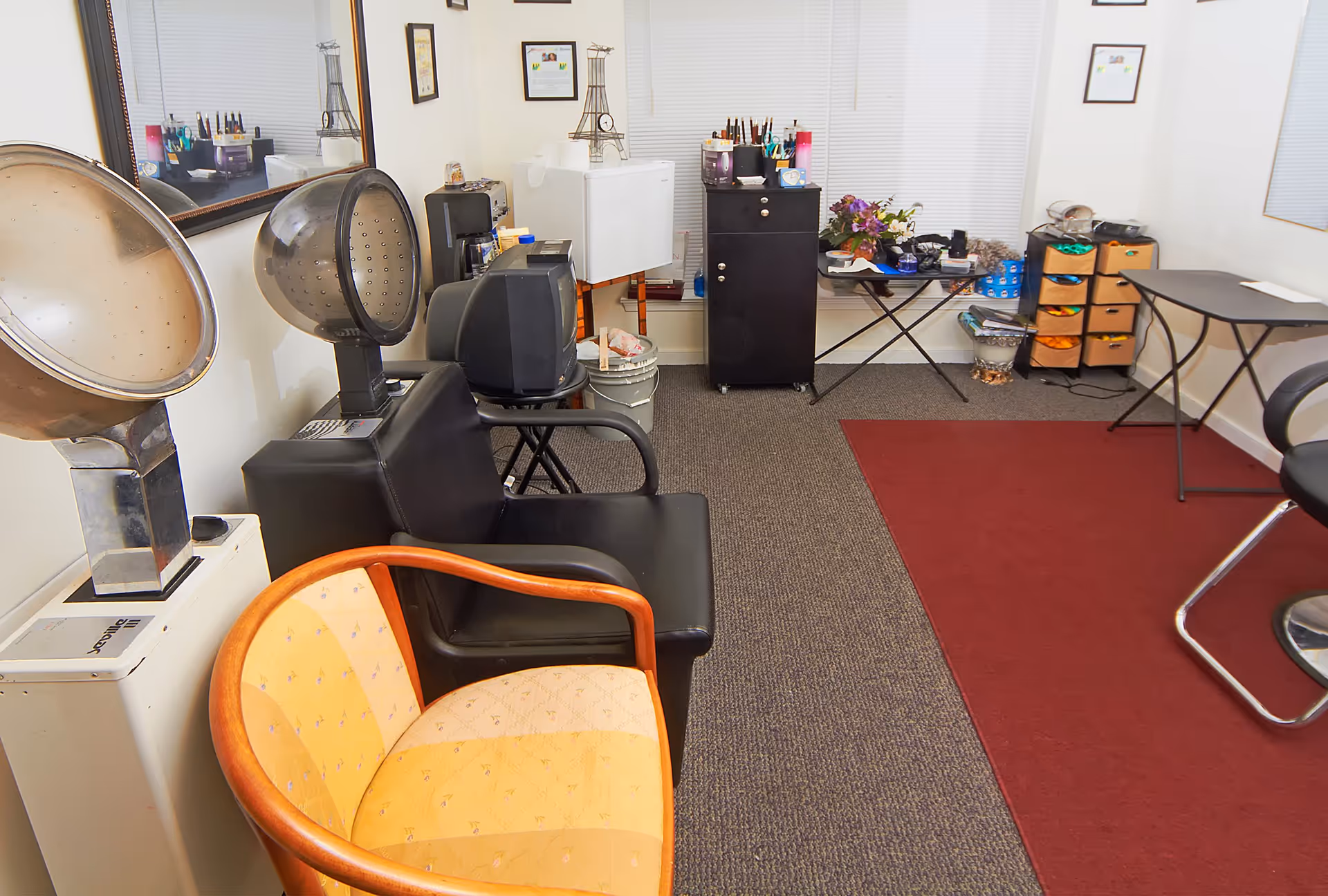 Interior salon/styling room with hooded hair dryers, seating, storage cabinets and a red area rug.