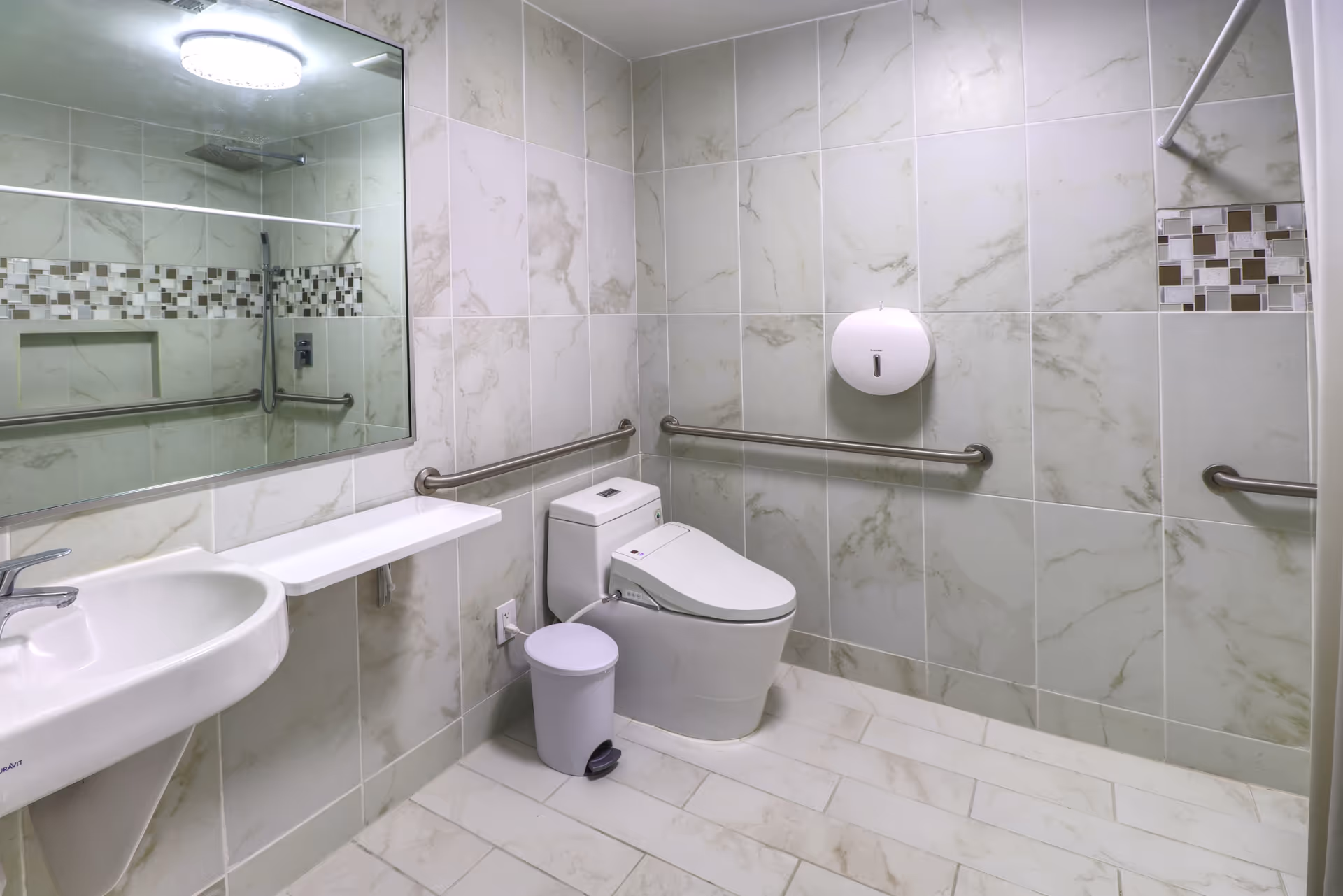 Accessible tiled bathroom with a toilet, sink, large mirror, grab bars and a shower area reflected in the mirror.