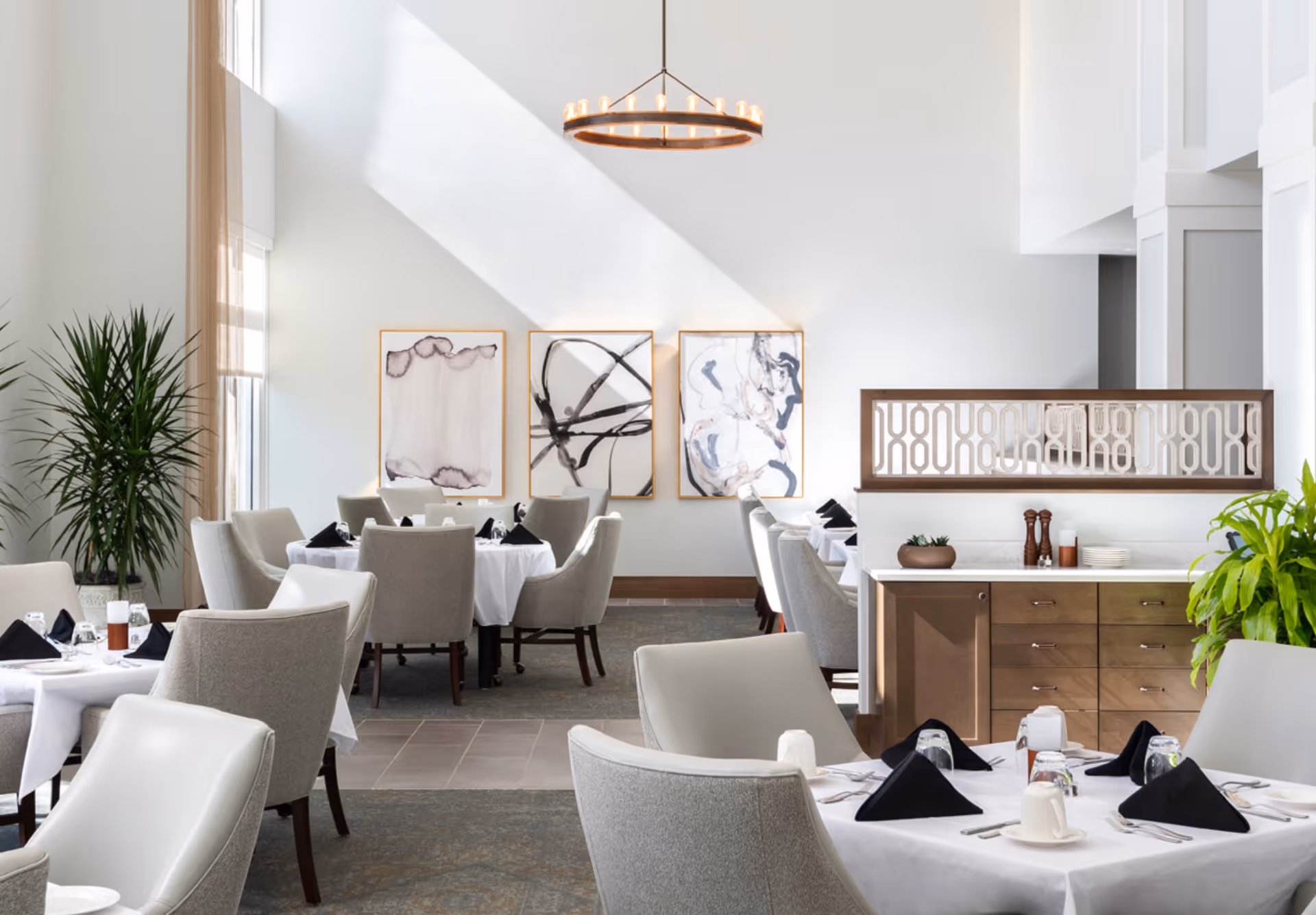 Bright, modern dining room with tables set with white linens and black napkins, upholstered chairs, artwork on the wall, and potted plants.
