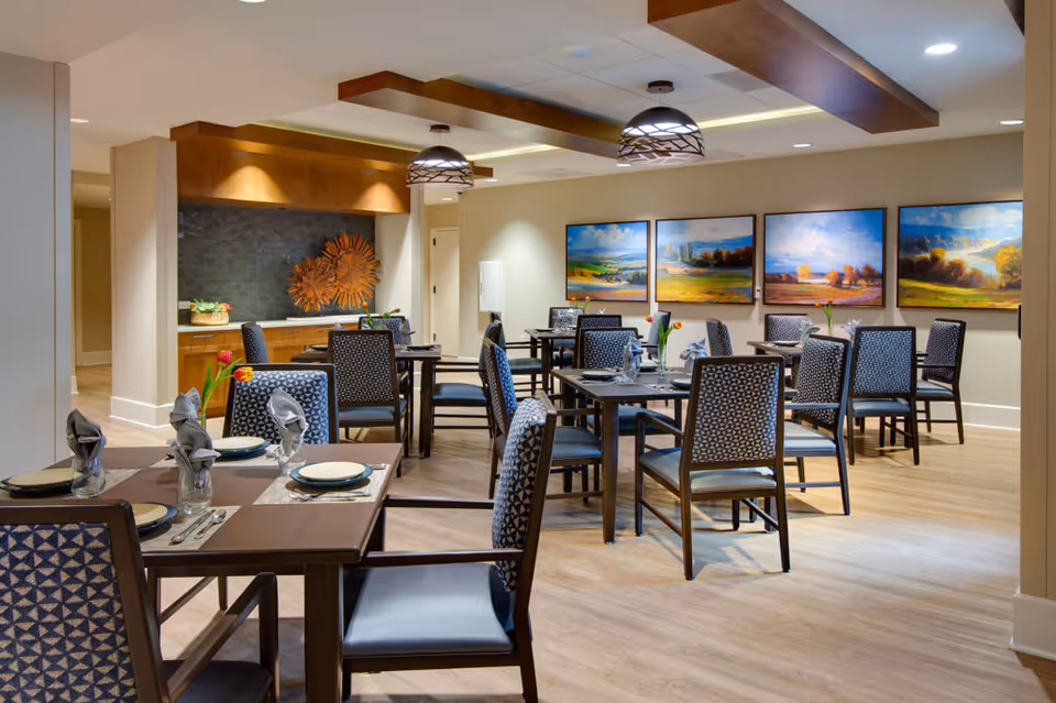 Well-lit dining room with multiple set tables and patterned chairs, landscape paintings on the wall, and decorative pendant lighting.