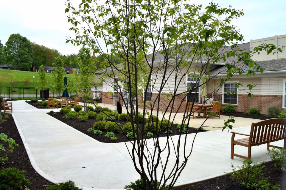 Outdoor patio area at Danbury Wooster featuring concrete walkways, wooden benches, small trees, and landscaped garden beds with shrubs. The building exterior has brick and siding with windows and patio tables with umbrellas.