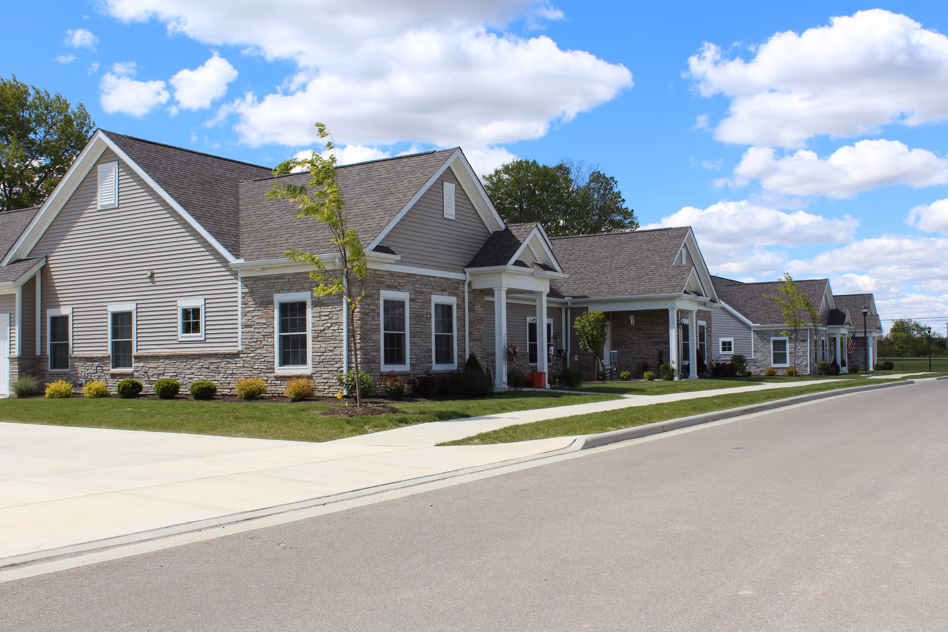 Row of single-story residential-style buildings with gray siding, stone accents, porches, and landscaped lawns along a street under a blue sky.
