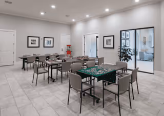 A spacious, well-lit room with multiple tables and chairs arranged for group activities or dining. One table has a partially completed puzzle on it. The walls are light-colored with framed artwork, and there is a large glass door leading to another room with more seating and plants.