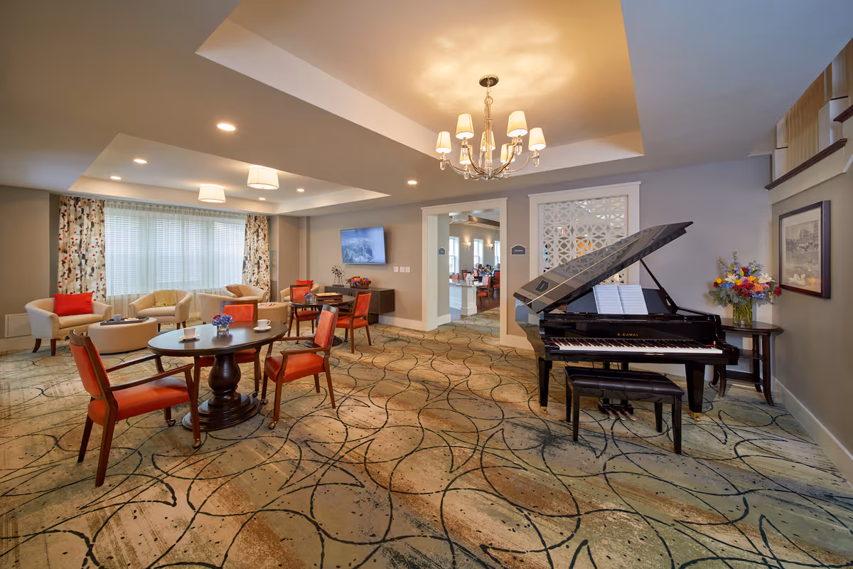 A spacious senior living common area with a grand piano on the right, a round table with four red chairs in the center, and a seating area with armchairs and a coffee table near a large window with patterned curtains. The room has a patterned carpet, a chandelier, and a wall-mounted TV.
