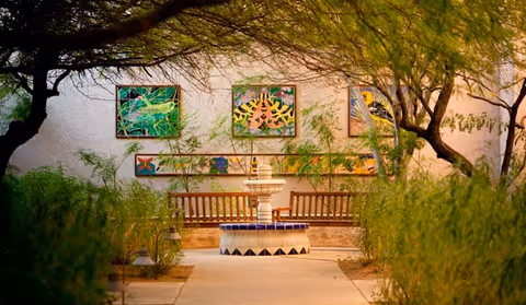 Outdoor courtyard area with a central water fountain surrounded by benches. The walls behind the benches display colorful framed artwork. Trees and greenery frame the pathway leading to the fountain.